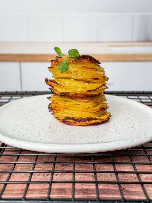 A neatly stacked tower of greek lemon potato stacks, thinly sliced and roasted, garnished with a small green herb, served on a white plate with a speckled pattern, placed on a black cooling rack.