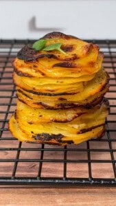 A stack of thinly sliced, golden-brown Greek lemon potato stacks sits on a black cooling rack, garnished with a small green herb, against a blurred background.