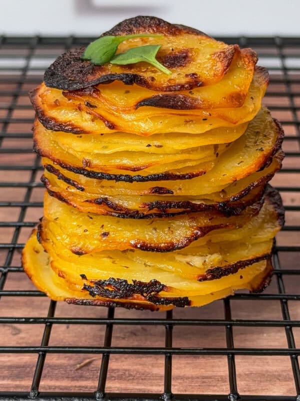 A stack of thinly sliced, golden-brown Greek lemon potato stacks sits on a black cooling rack, garnished with a small green herb, against a blurred background.
