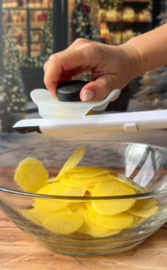 A hand uses a mandoline slicer to cut yellow potatoes into thin slices for Greek lemon potato stacks over a glass bowl on a wooden surface, with a festive, blurred background.