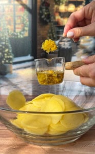 A hand pours olive oil and grated lemon zest from a measuring cup onto sliced potatoes in a glass bowl, preparing festive greek lemon potato stacks, with a wintery outdoor scene visible through the window in the background.