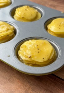 Close-up of a muffin tin filled with greek lemon potato stacks—thinly sliced potatoes seasoned with herbs and spices, ready to be baked. The tin rests on a wooden surface.