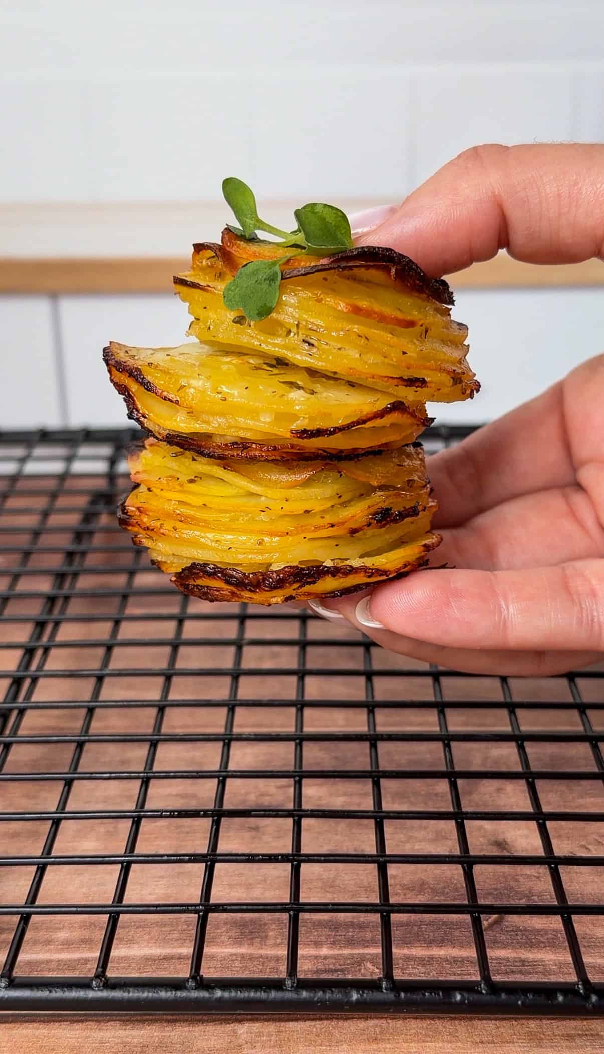 A hand holds a stack of crispy, golden greek lemon potato stacks, thinly sliced and garnished with a sprig of green herb, above a black cooling rack on a wooden surface. - 4