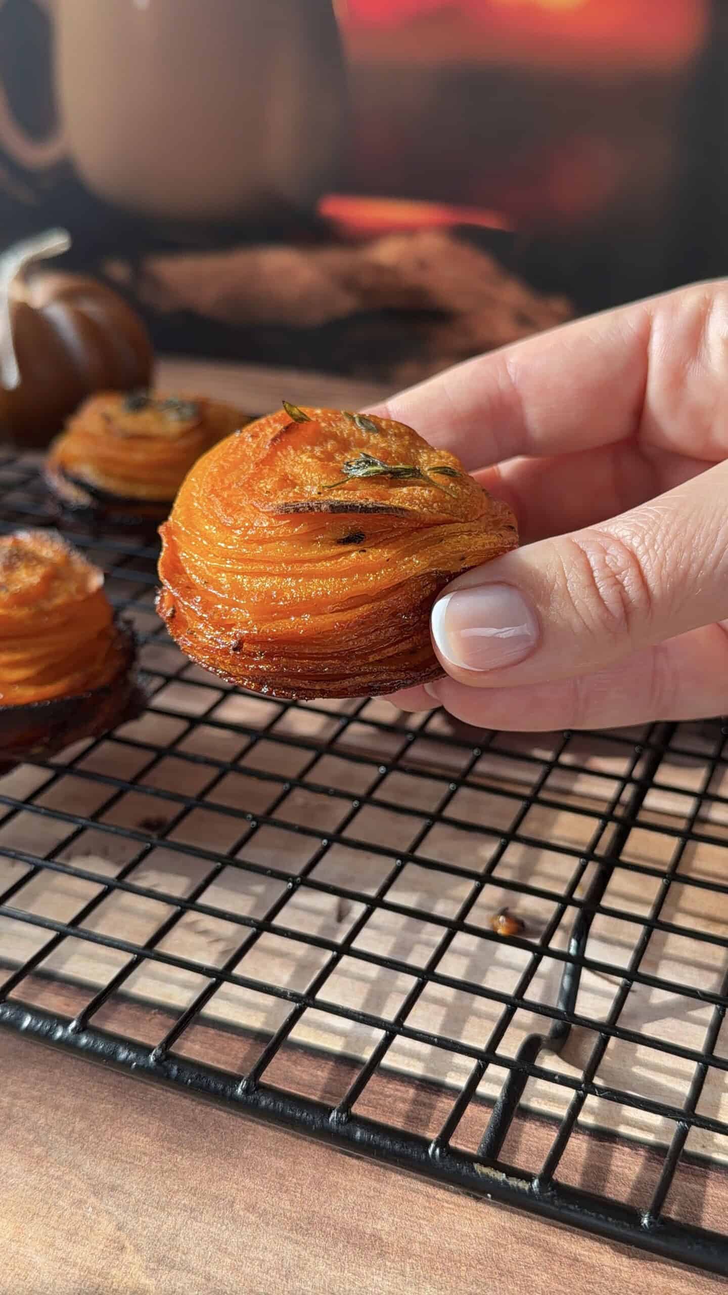 A hand holds a small, spiral-shaped Sweet Potato Stack on a cooling rack, with more stacks and a blurred background visible. - 4