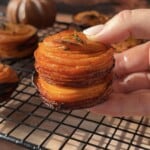 A hand holds a stack of thinly sliced, roasted sweet potato stacks with crispy edges, garnished with herbs, over a cooling rack. Mini pumpkins and a cup are softly blurred in the background.