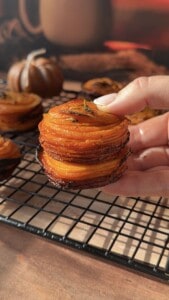 A hand holds a stack of thinly sliced, roasted sweet potato stacks with crispy edges, garnished with herbs, over a cooling rack. Mini pumpkins and a cup are softly blurred in the background.