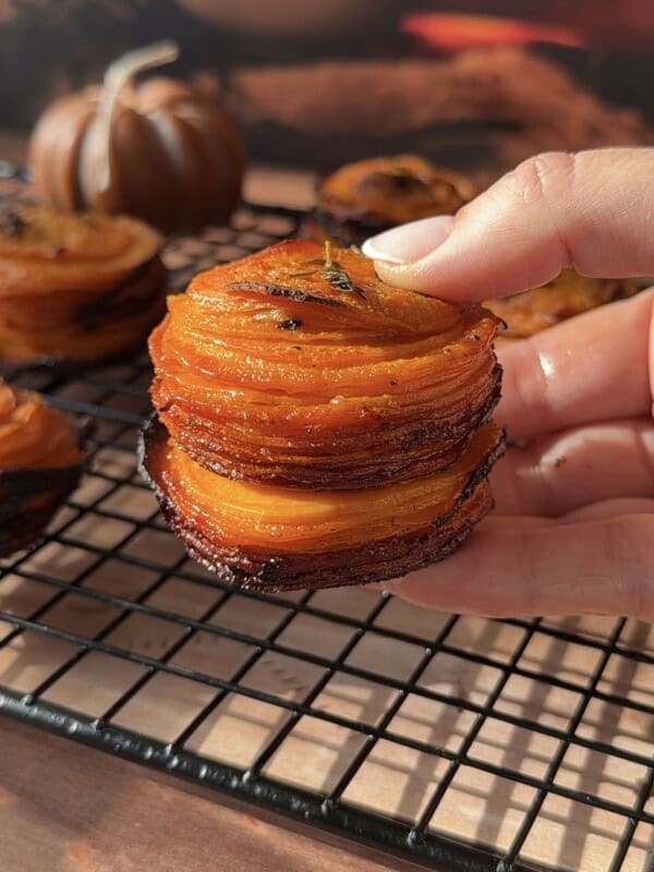 A hand holds a stack of thinly sliced, roasted sweet potato stacks with crispy edges, garnished with herbs, over a cooling rack. Mini pumpkins and a cup are softly blurred in the background.