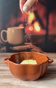 A hand sprinkles salt over a brown dish of Sweet Potato Stacks garnished with a slice of lemon, while a blurred background reveals a mug and the warm glow of a fireplace.