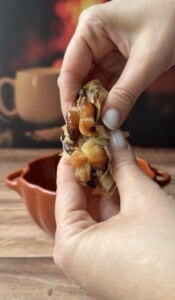 A close-up of hands squeezing roasted garlic cloves from the bulb over a brown bowl, with a blurred mug and warm background—perfect for flavoring Sweet Potato Stacks.