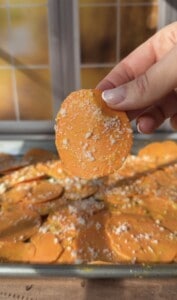 A hand holds up a round, orange sweet potato slice sprinkled with grated cheese, with more Sweet Potato Stacks and cheese spread out on a baking sheet in the background. Sunlight streams through a window behind.