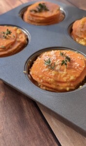 Close-up of a muffin tin filled with Sweet Potato Stacks—layers of sliced sweet potatoes topped with herbs and grated cheese—ready for baking on a wooden surface.