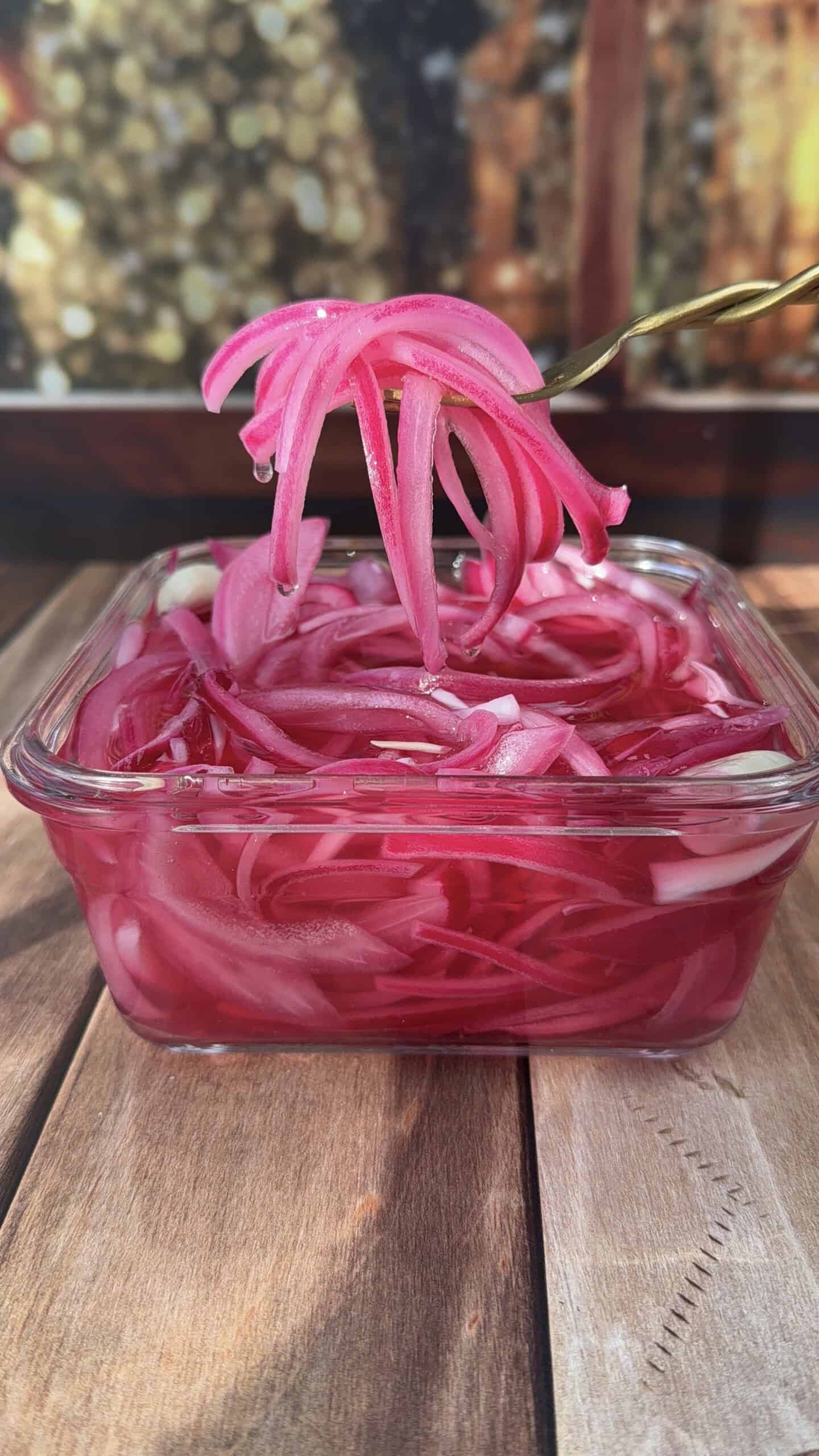 A glass container filled with bright pink pickled red onions sits on a wooden surface, perfect as a topping for Smoked Salmon Jerky. A fork lifts a portion of the onions above the jar, while the background is softly blurred.