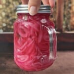 A hand holds a glass jar filled with bright pink pickled onions in a liquid, set against a blurred, warm-toned background with lights and a wooden surface—perfect as a tangy topping for smoked salmon jerky.