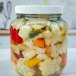 A large glass jar filled with quick pickled veggies—cauliflower, carrots, and peppers—sits on a white countertop in a bright kitchen setting.