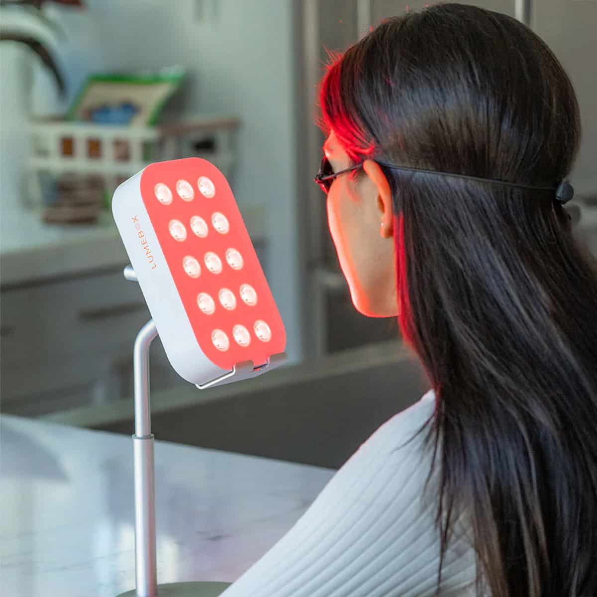 A woman with long dark hair wears protective goggles while sitting in front of a tabletop red light therapy device emitting bright red lights in a kitchen setting, enjoying her self-care routine and looking out for exclusive deals.