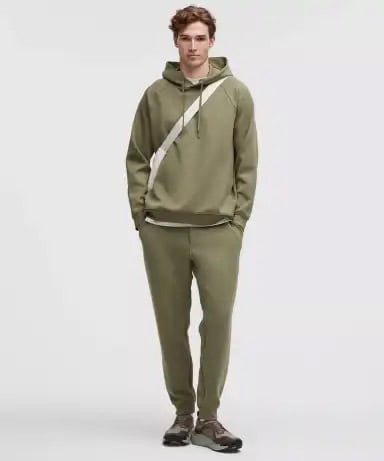 A person stands facing forward, wearing an olive green hooded sweatshirt, matching jogger pants, dark sneakers, and a light-colored crossbody bag against a plain light backgroundโperfect for shopping exclusive deals in style.
