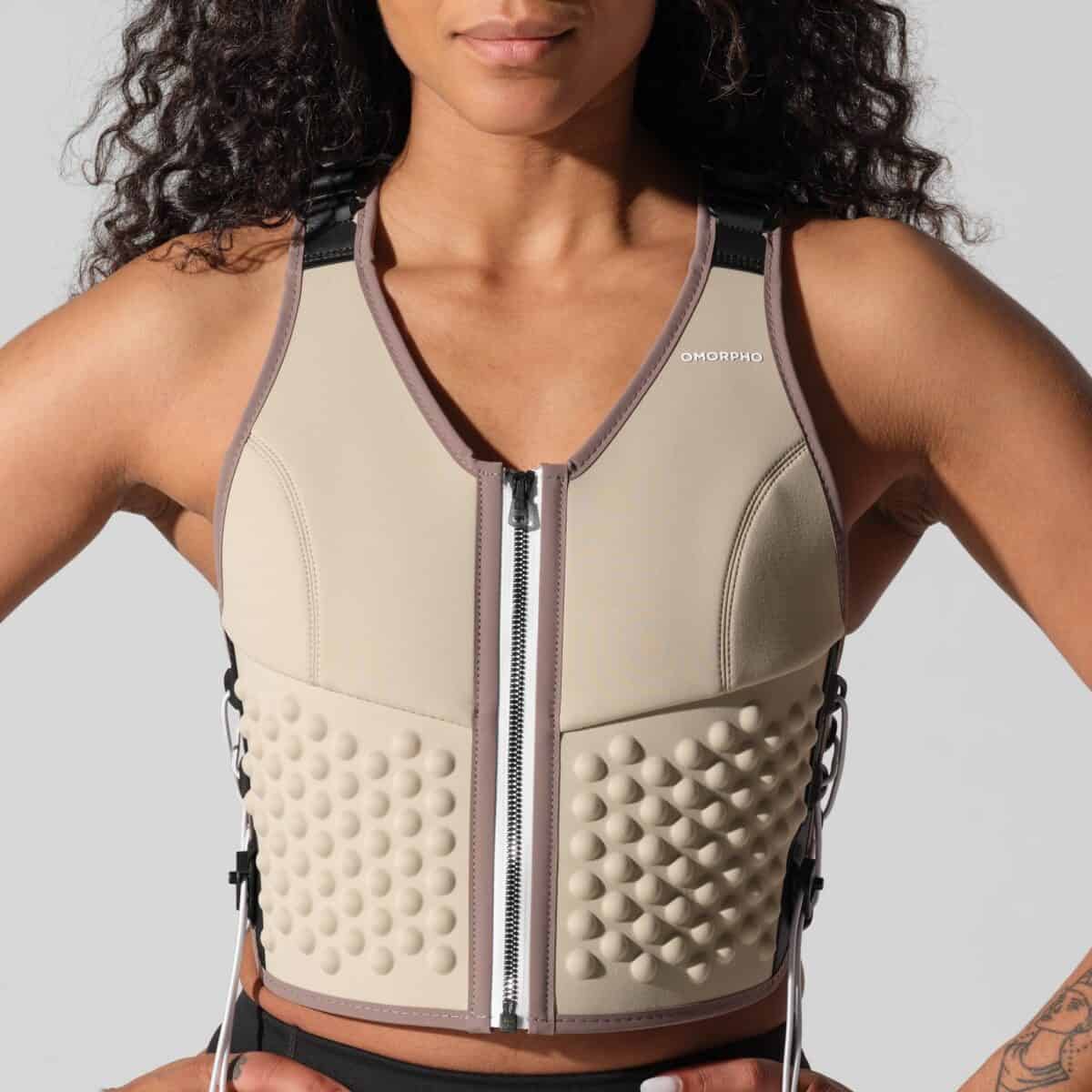 A woman wears a beige, textured weighted vest with a front zipper and black adjustable straps. Featuring raised circular bumps and the "OMORPHO" logo on the left chest, this vest is available now with exclusive deals.