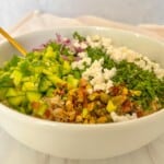 A white bowl filled with chopped green bell peppers, red onions, crumbled cheese, fresh herbs, pistachios, crushed red pepper flakes, and grains, with a gold-colored spoon resting inside.