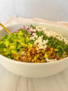 A white bowl filled with chopped green bell peppers, red onions, crumbled cheese, fresh herbs, pistachios, crushed red pepper flakes, and grains, with a gold-colored spoon resting inside.