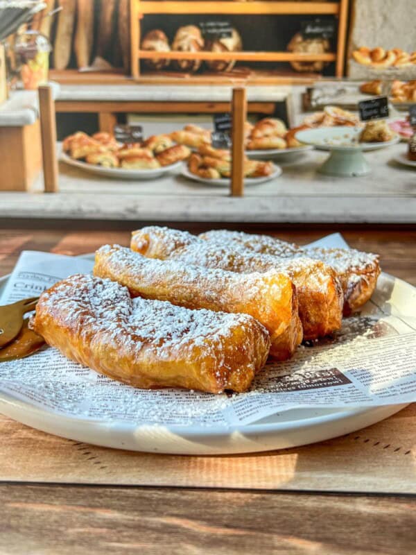A plate of golden, flaky pastries dusted with powdered sugar sits on a newspaper-lined dish, with more baked goods and pastries visible in the blurred background of a bakery display.