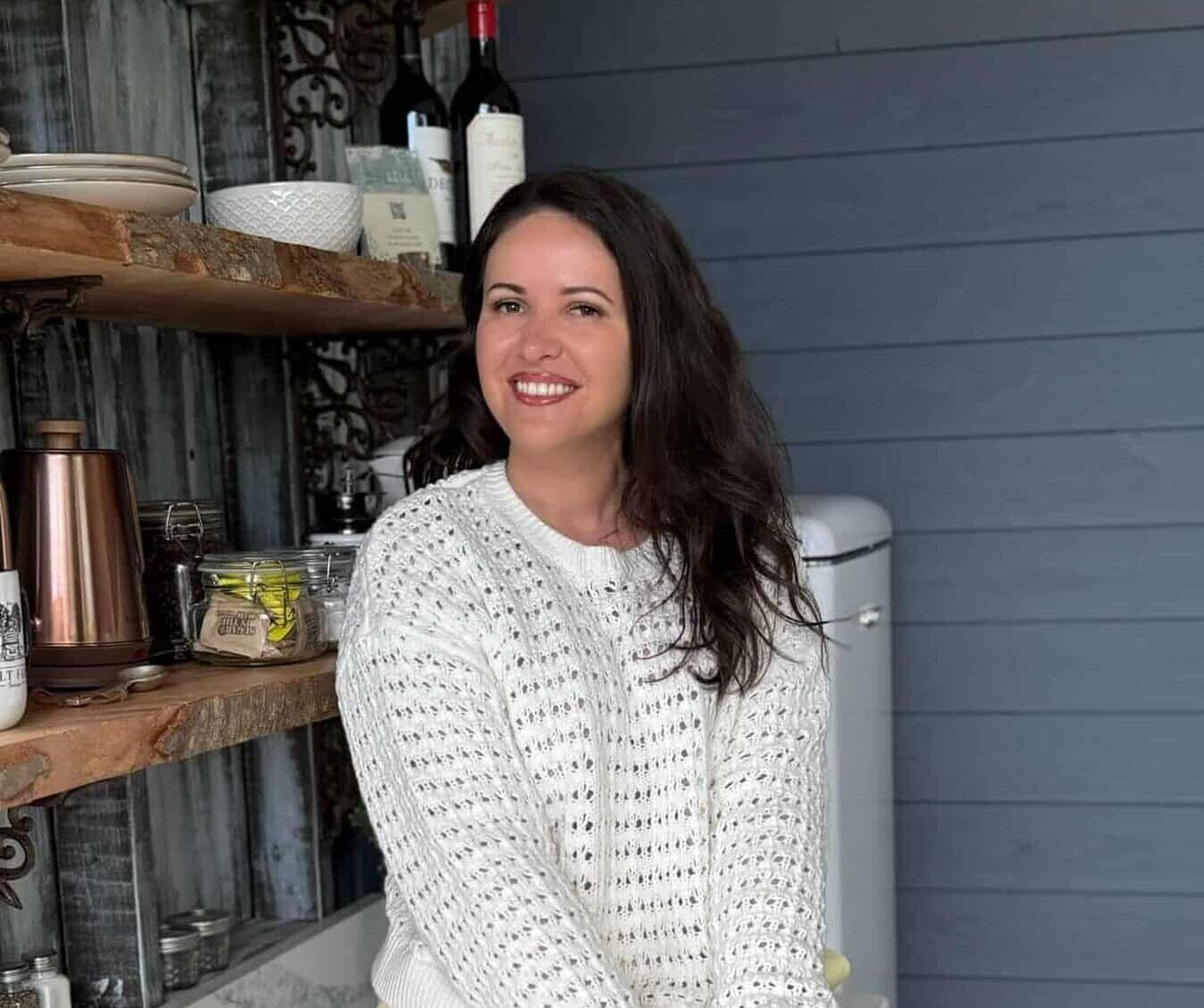 A woman with long brown hair, wearing a white knit sweater and yellow shorts, sits smiling on a marble countertop in a cozy kitchen with wooden shelves, dishware, and a vintage refrigerator.