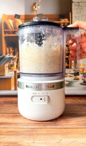A hand holds a KitchenAid food processor filled with a creamy, herb-speckled mixture, perfect for pairing with honey glazed carrots, on a wooden countertop in a cozy kitchen setting.