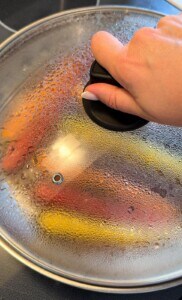 A hand holds the handle of a glass pot lid covered in condensation, with colorful vegetables like honey glazed carrots, red, and yellow peppers steaming inside the pot on a stovetop.