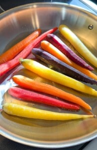 Several whole carrots in a variety of colors—orange, yellow, red, and purple—are arranged in a single layer inside a stainless steel pan, ready to be cooked into delicious Honey Glazed Carrots.