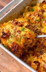 A close-up of a casserole dish filled with cheesy baked casserole. A serving is being lifted out with a spoon, showing layers of melted cheese, ground meat, and herbs, garnished with fresh parsley.