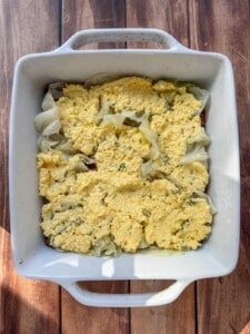 A white square baking dish containing a layer of cooked cabbage topped with an uneven layer of yellow, crumbly cornbread mixture, sitting on a wooden surface.