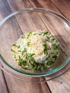 A glass bowl filled with ricotta cheese, grated parmesan, and chopped fresh basil, sitting on a wooden surface.