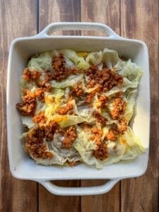 A white baking dish filled with cabbage rolls topped with a chunky tomato and meat sauce, set on a wooden surface.