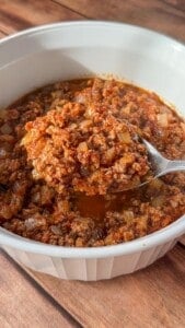 A close-up of a spoonful of ground meat in a rich red sauce with diced onions, held over a white bowl filled with the same mixture, on a wooden surface.