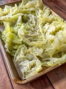 A baking sheet covered with wilted, pale green cabbage leaves spread out in a single layer on a wooden surface.