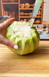 A hand holds a green cabbage on a wooden cutting board while a knife cuts around its core. Shelves with bread and baked goods are visible in the blurred background.