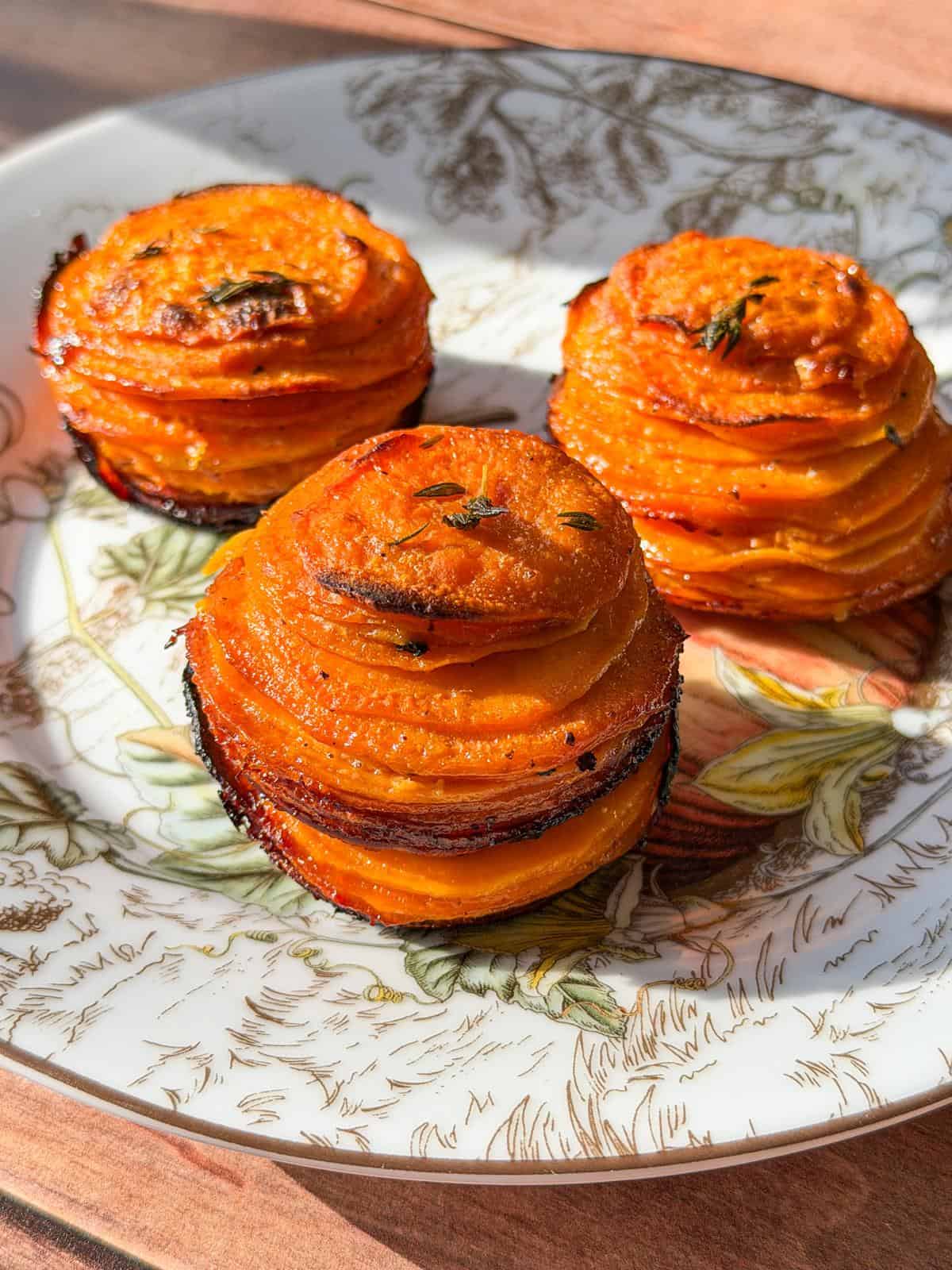 Three stacks of thinly sliced, roasted sweet potatoes, browned at the edges, are arranged on a decorative plate with a botanical pattern. Sunlight highlights their golden-orange color and crisp texture. - 3