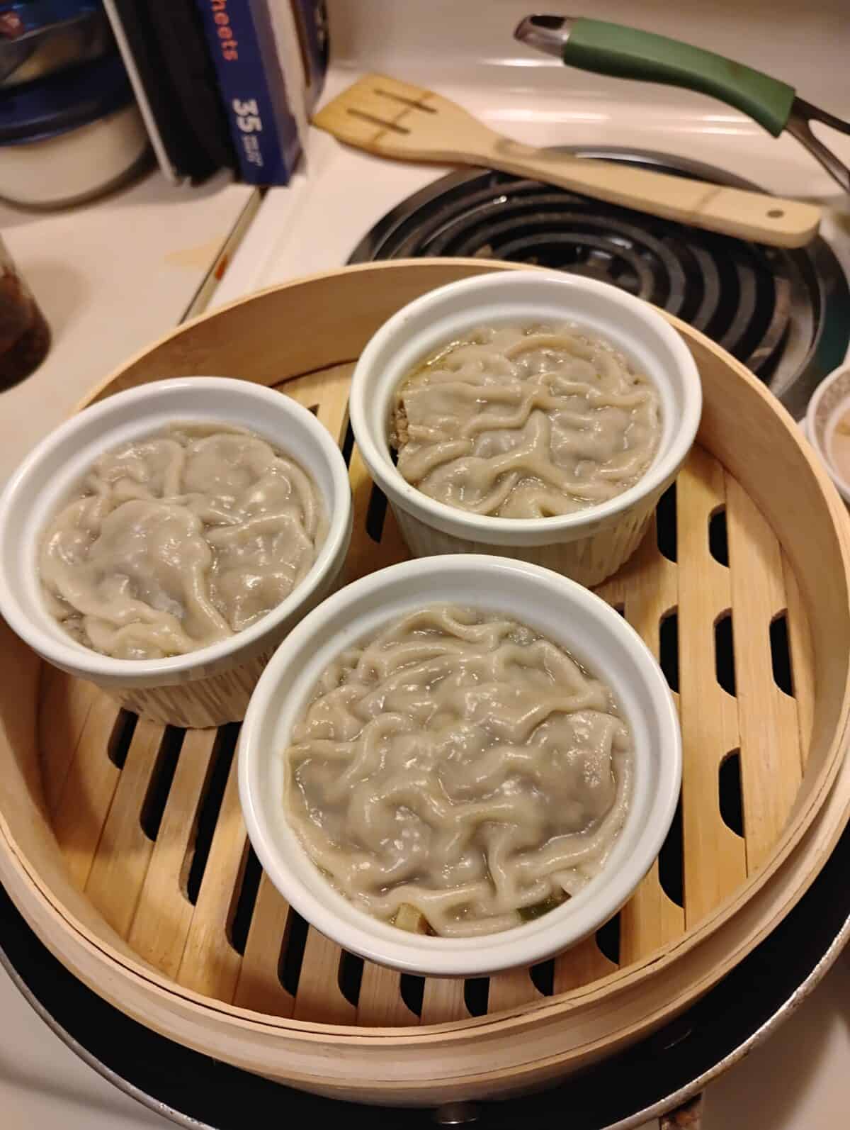 Three small white ramekins filled with a light brown, wrinkled mixture, reminiscent of VIRAL PORK DUMPLING LASAGNA, are steaming inside a bamboo steamer basket on a stovetop. A green-handled tool and various kitchen items are visible in the background.