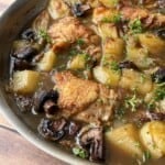 A skillet filled with golden-brown, crispy chicken thighs, potatoes, mushrooms, and fresh herbs in a savory broth, all cooked together and garnished with chopped parsley—a delicious crispy chicken thighs recipe.