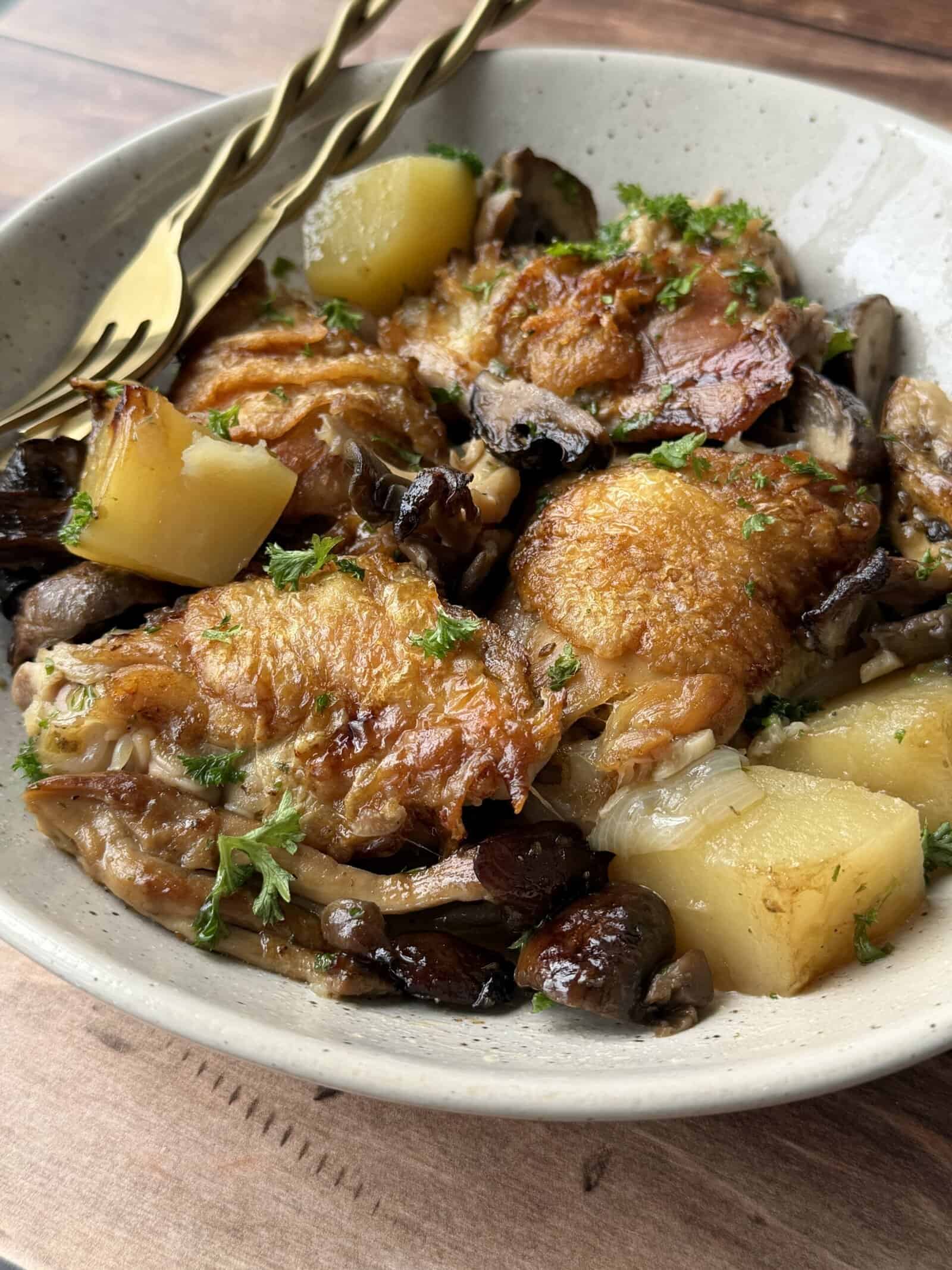 A bowl of braised crispy chicken thighs recipe with golden skin, potatoes, mushrooms, and fresh parsley garnish, served with a pair of golden utensils on a wooden surface.
