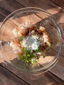 A glass bowl containing chopped shrimp, green onions, a heap of white flour, and seasoning, placed on a wooden surface in natural sunlight.