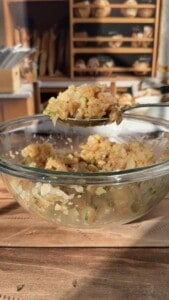 A glass bowl filled with a rice and vegetable mixture sits on a wooden table. A spoon holding a portion of the mixture is raised above the bowl. Shelves with bread loaves are visible in the blurred background.
