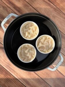 Three white ramekins filled with uncooked, wavy dough are arranged in a round black Staub pot with water, set on a wooden surface. Sunlight casts shadows on the pot and wood.