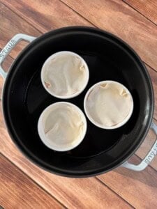 Three white ramekins covered with dough sit in a black round baking dish filled with water, placed on a wooden surface.