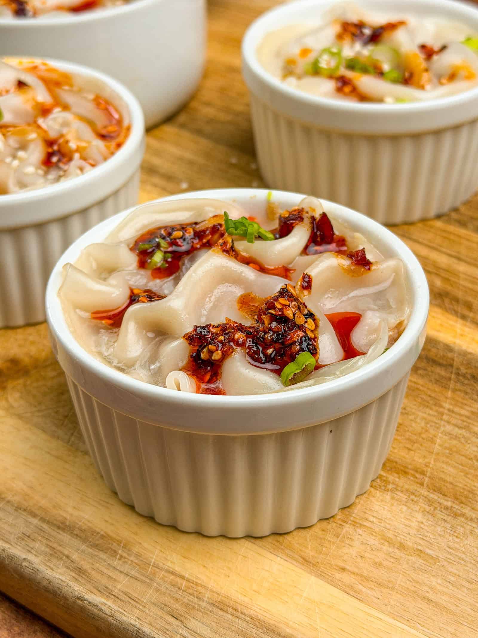 A small white ramekin filled with wide, wavy rice noodles topped with chili oil and herbs sits on a wooden surface, inspired by the flavors of VIRAL PORK DUMPLING LASAGNA. More ramekins with this dish appear in the background.