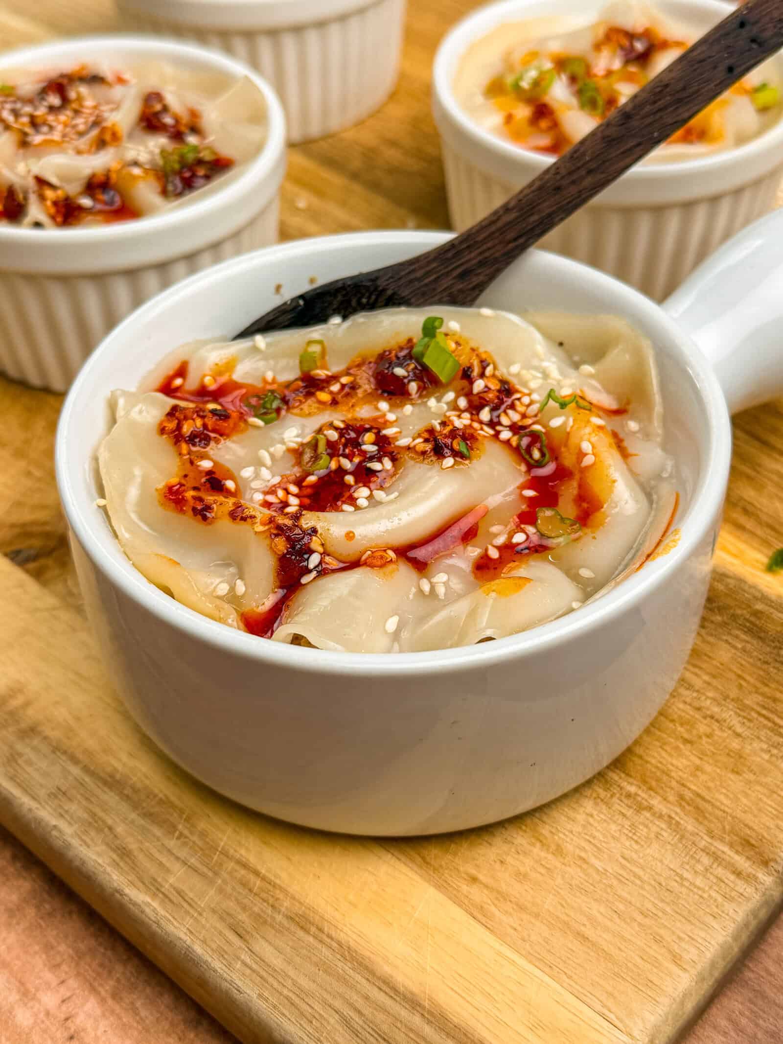 A white ramekin filled with chili oil dumplings, inspired by the VIRAL PORK DUMPLING LASAGNA trend, is garnished with sesame seeds and green onions on a wooden board. A dark spoon rests on the edge, with more dumplings in the background.