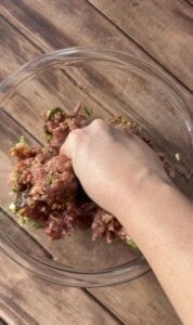 A hand mixes ground meat with chopped green herbs and seasonings in a clear glass bowl on a wooden surface.