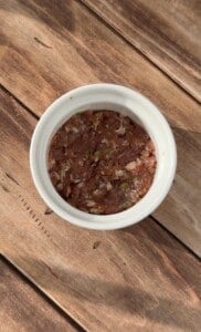 A white bowl filled with a mixture of minced meat and herbs sits on a wooden surface, lit by natural sunlight.