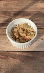 A white ramekin filled with chopped shrimp mixed with green herbs and seasonings sits on a wooden surface, illuminated by sunlight.