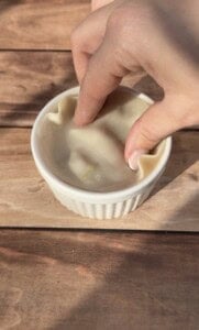 A hand presses a sheet of dough into a small white ramekin on a wooden surface, shaping the dough to fit the dish.