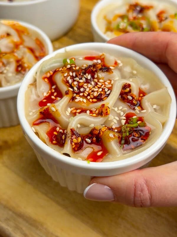 A hand holds a white ramekin filled with flat noodles topped with chili oil, sesame seeds, and sliced green onions. Three more similar noodle dishes are in the background on a wooden surface.